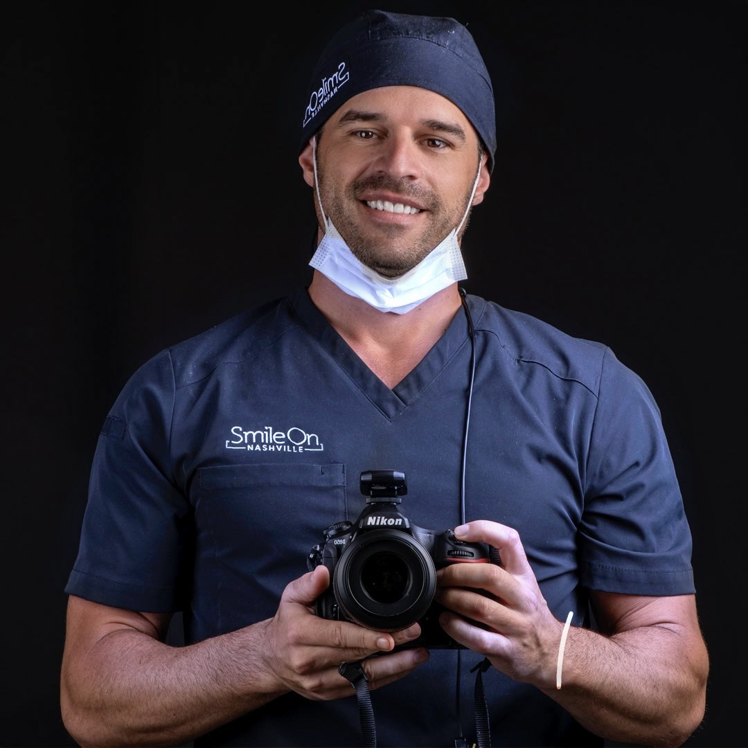 Dr. Jeff Trembley smiling in his Smile On Nashville scrub cap, holding his studio camera against a black backdrop